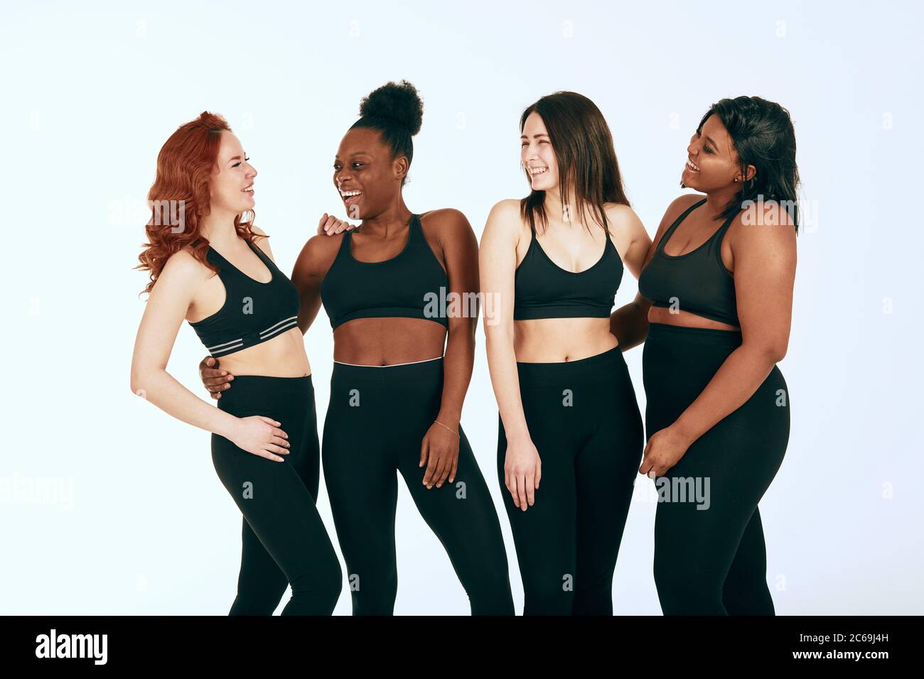 de cuatro mujeres de diferentes razas, tipos y tamaños en el traje deportivo hablando animado y riendo sobre fondo Fotografía de stock -