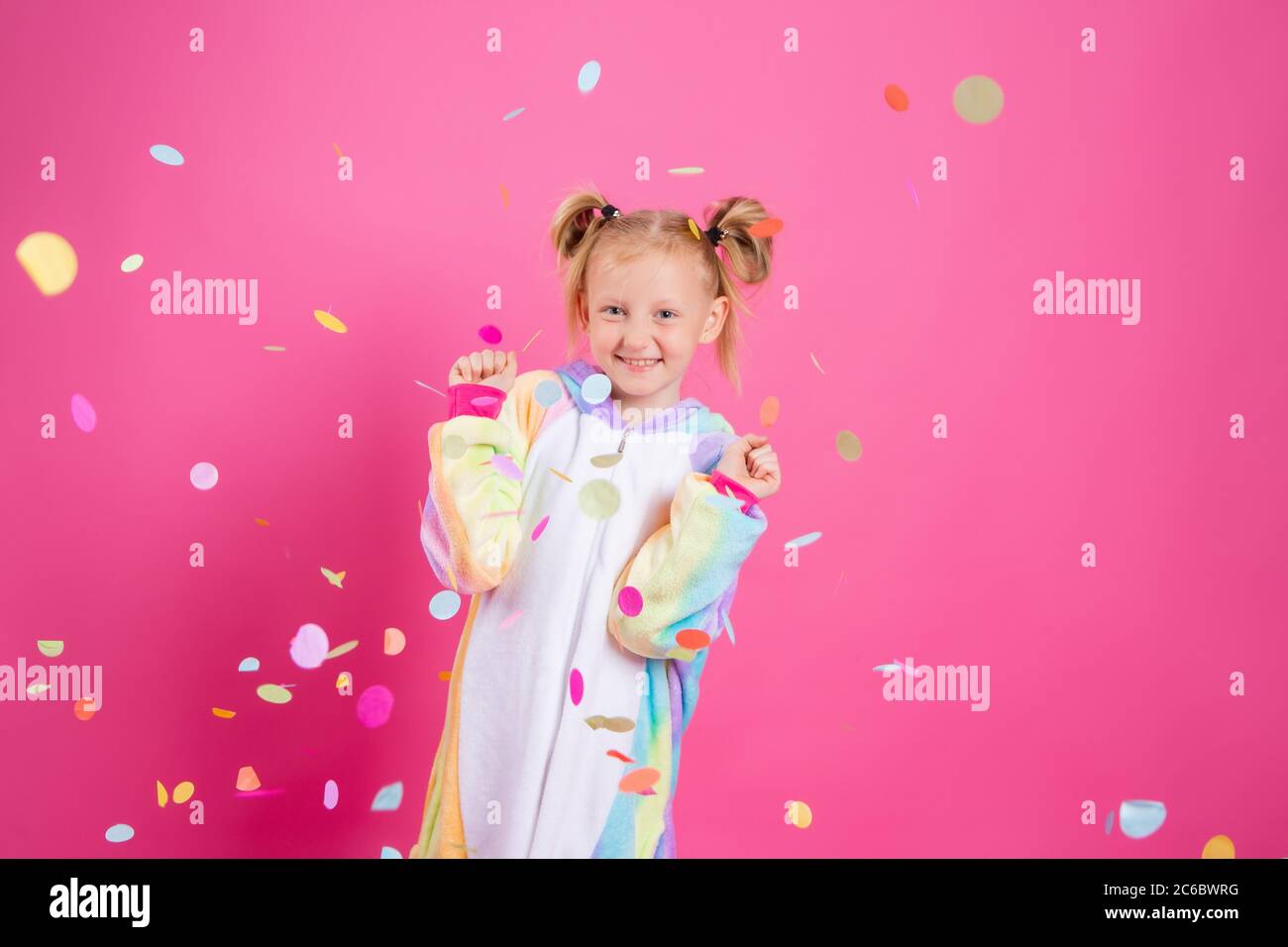 Feliz niña en kigurumi unicorn sobre un fondo rosa se regocija en confeti multicolor, espacio para el texto Foto de stock