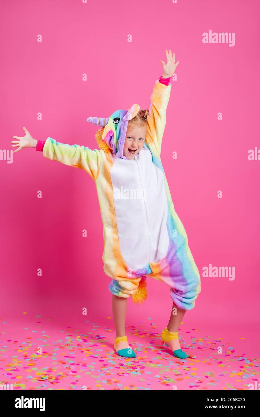 Feliz niña en kigurumi unicorn sobre un fondo rosa se regocija en confeti multicolor, espacio para el texto Foto de stock