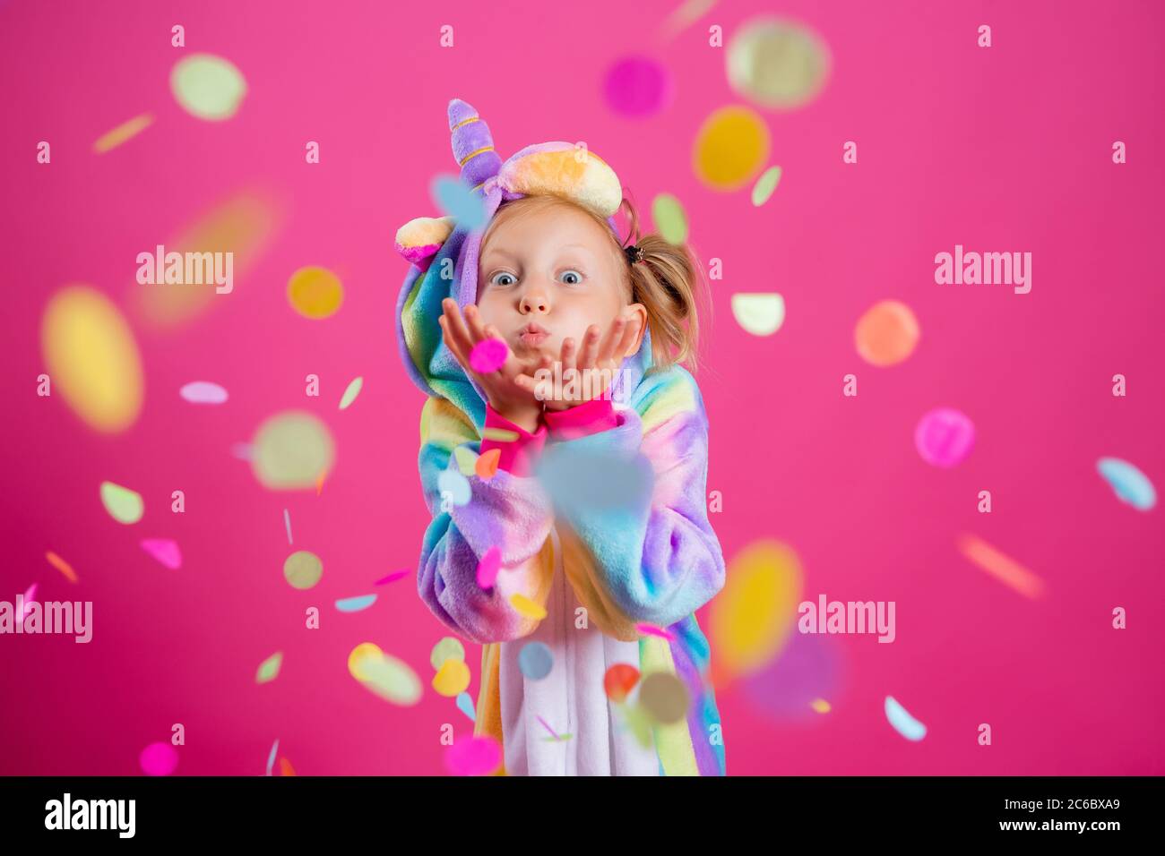 Feliz niña en kigurumi unicorn sobre un fondo rosa se regocija en confeti multicolor, espacio para el texto Foto de stock
