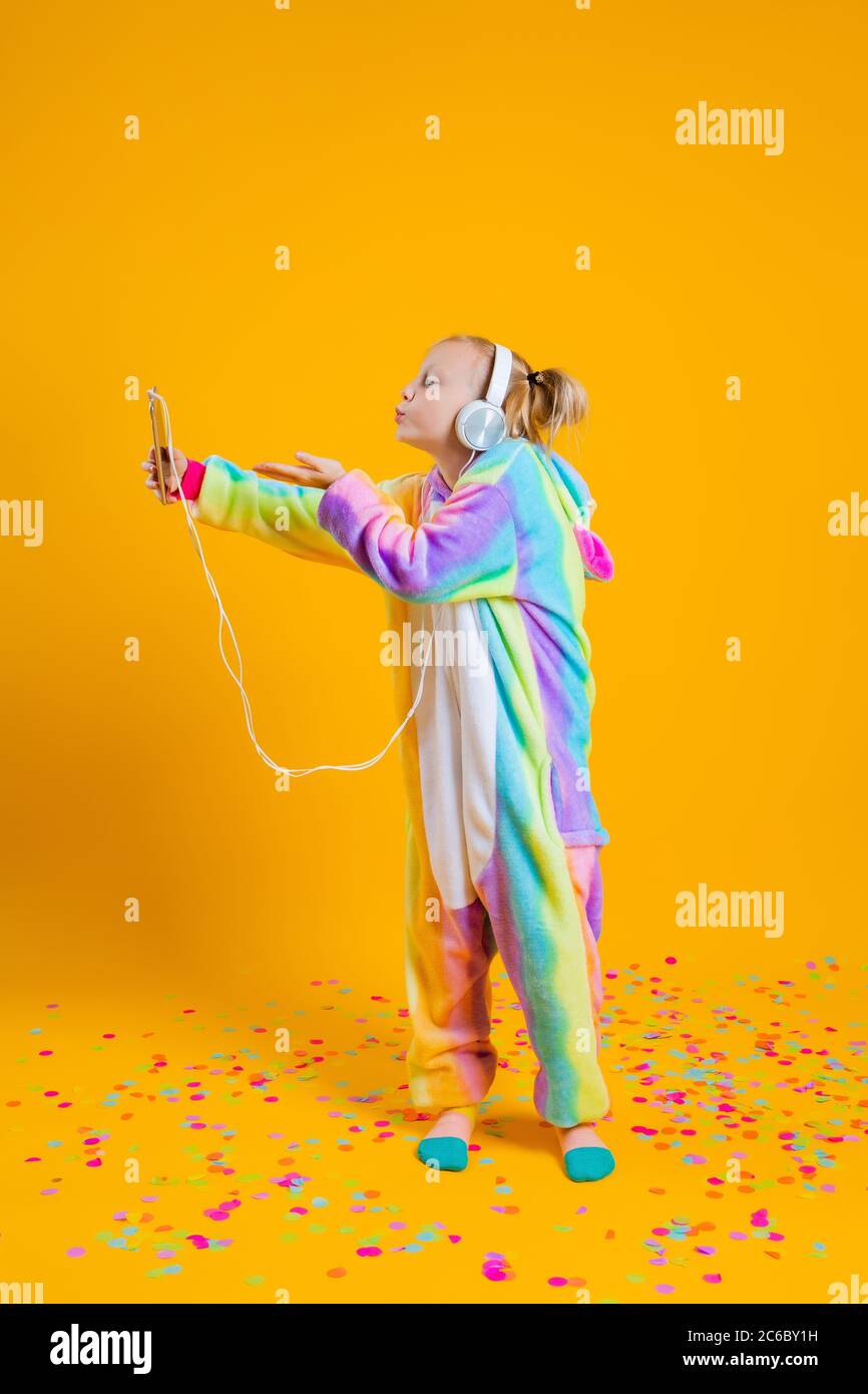 Feliz niña en kigurumi unicorn bailando sobre un fondo amarillo entre confeti multicolor Foto de stock