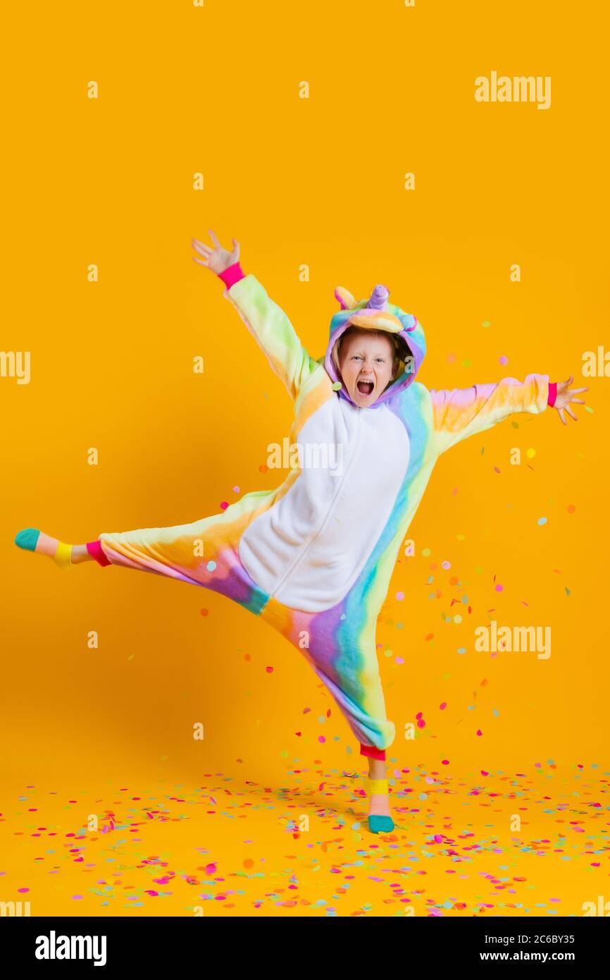 Feliz niña en kigurumi unicorn bailando sobre un fondo amarillo entre confeti multicolor Foto de stock