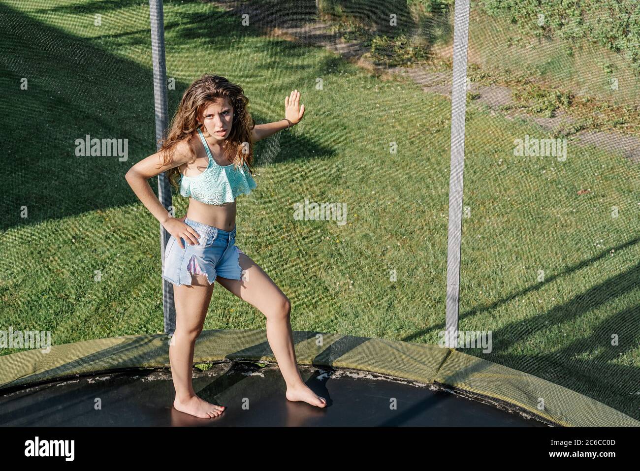 Foto horizontal de joven morena adolescente en un trampolín apoyado con su  mano en la red en un parque. La chica lleva pantalones cortos y una  camiseta y se ve Fotografía de