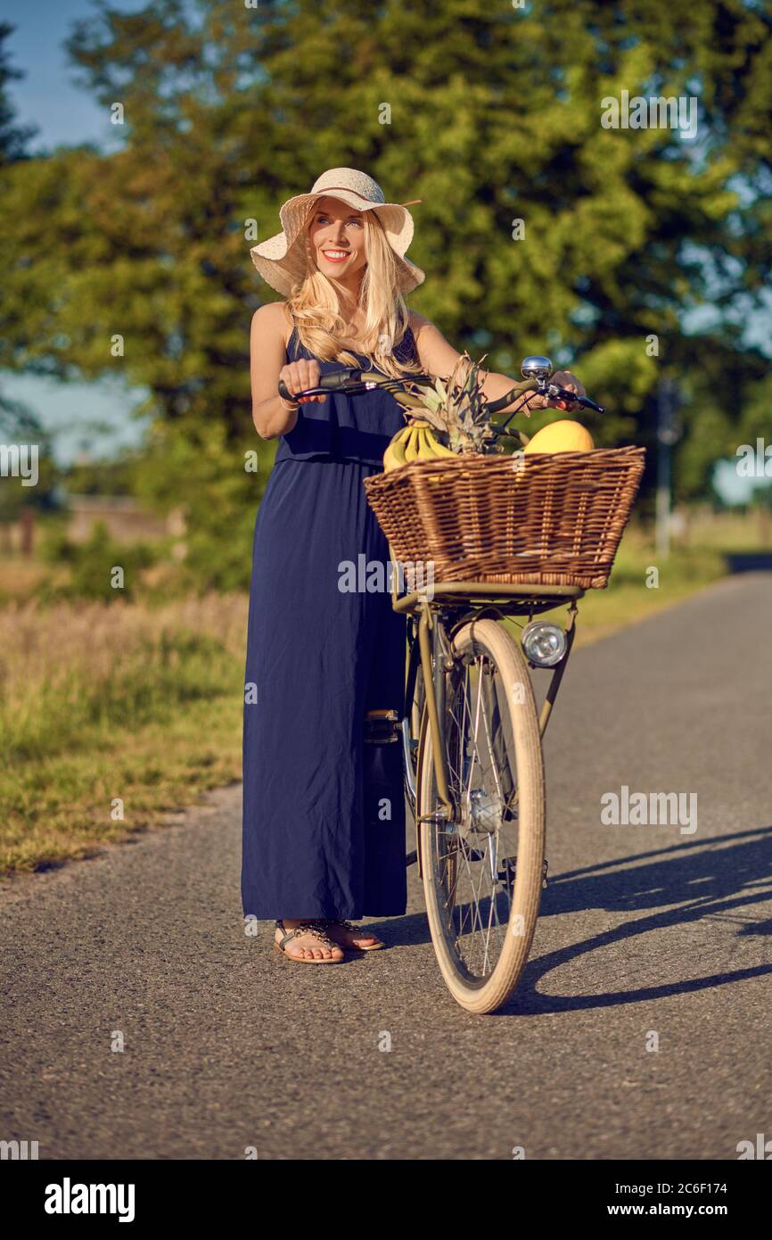 Mujer rubia atractiva en un amplio sombrero de paja brimmed que rueda una  bicicleta con productos frescos saludables en la cesta a lo largo de un  camino rural en el sol de