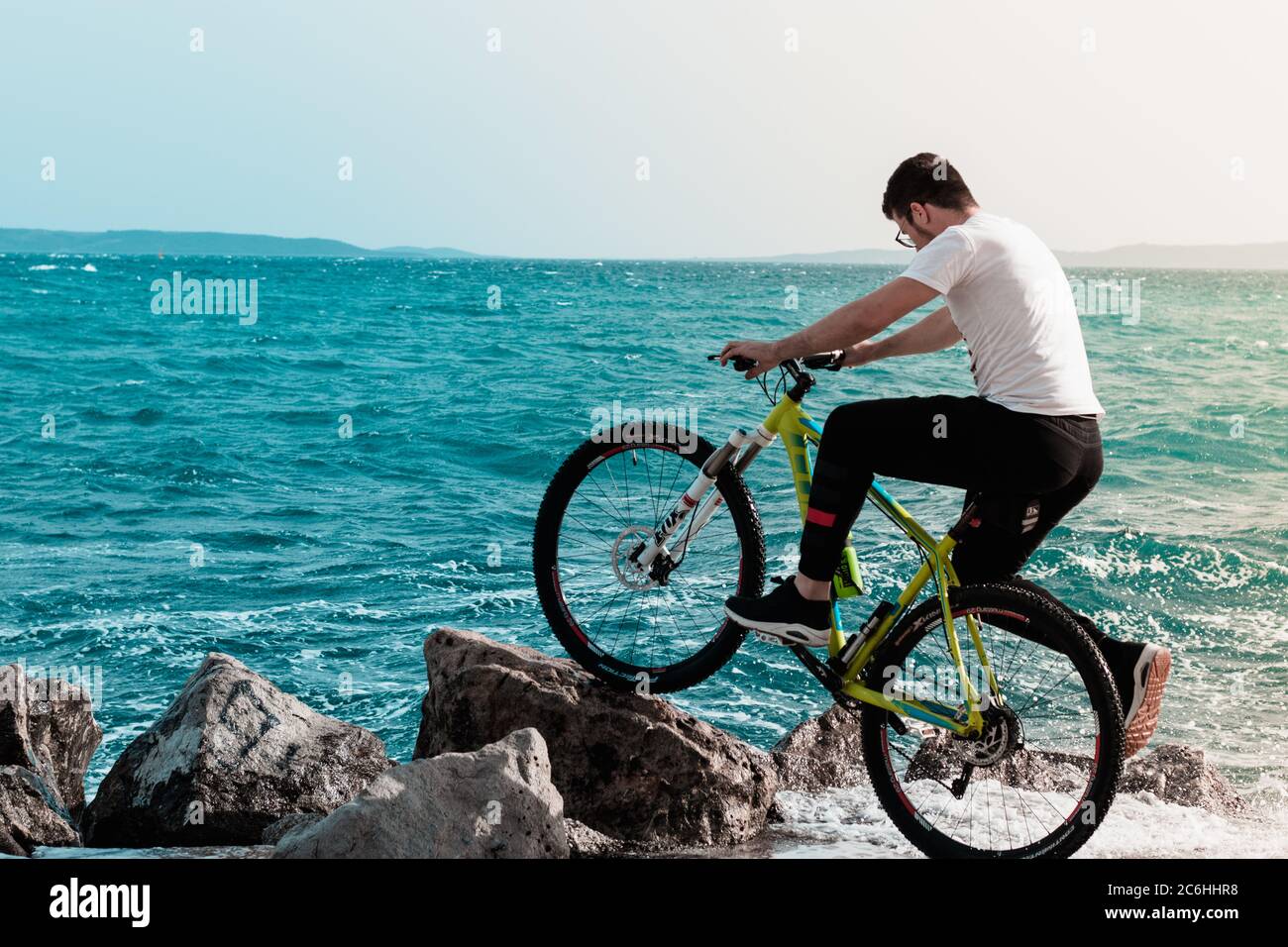 Split,Croacia Mayo 2020 joven de pie en la orilla con una bicicleta, se inclinó sobre una roca de playa. Olas golpeando la orilla en un día ventoso, salpicándolo Foto de stock