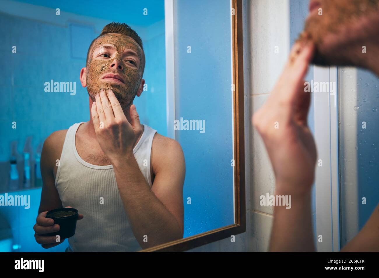 Joven aplicando máscara facial frente al espejo en el baño de la casa. Temas cuidado de la piel, tratamiento y belleza de los hombres. Foto de stock