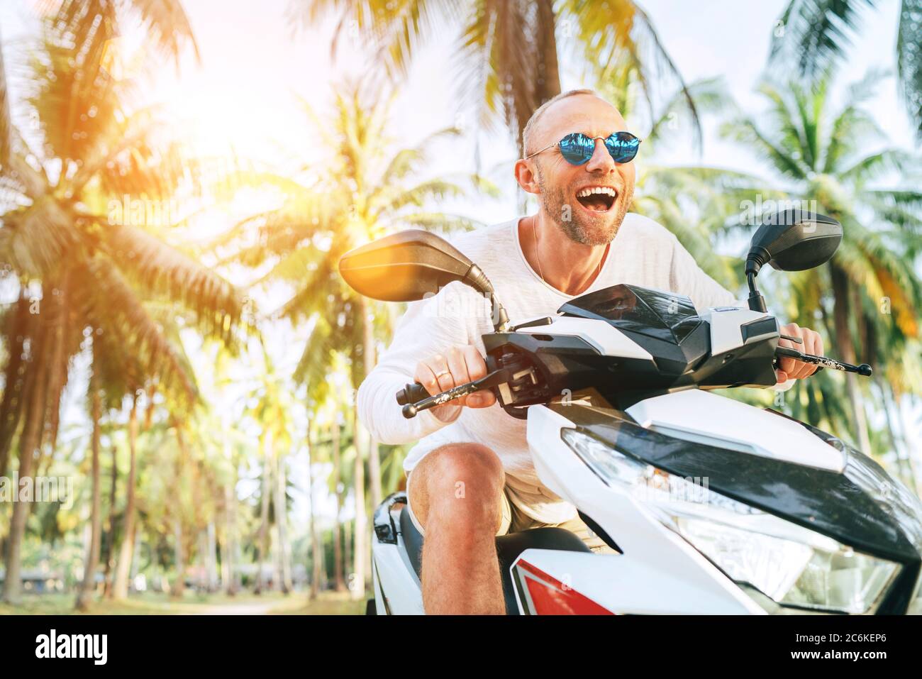 Feliz hombre sonriente turista en gafas de sol montar moto scooter durante  sus vacaciones tropicales bajo palmeras con rayos de sol brillantes  Fotografía de stock - Alamy
