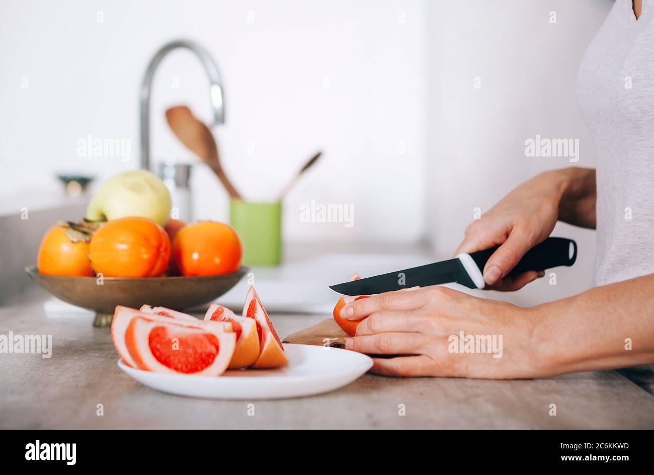 Las manos de la mujer joven cortando pomelo usando un cuchillo y una tabla de cortar en la cocina moderna imagen de primer plano. Un montón de manzanas, frutas de la fruta, kaki y orán Foto de stock
