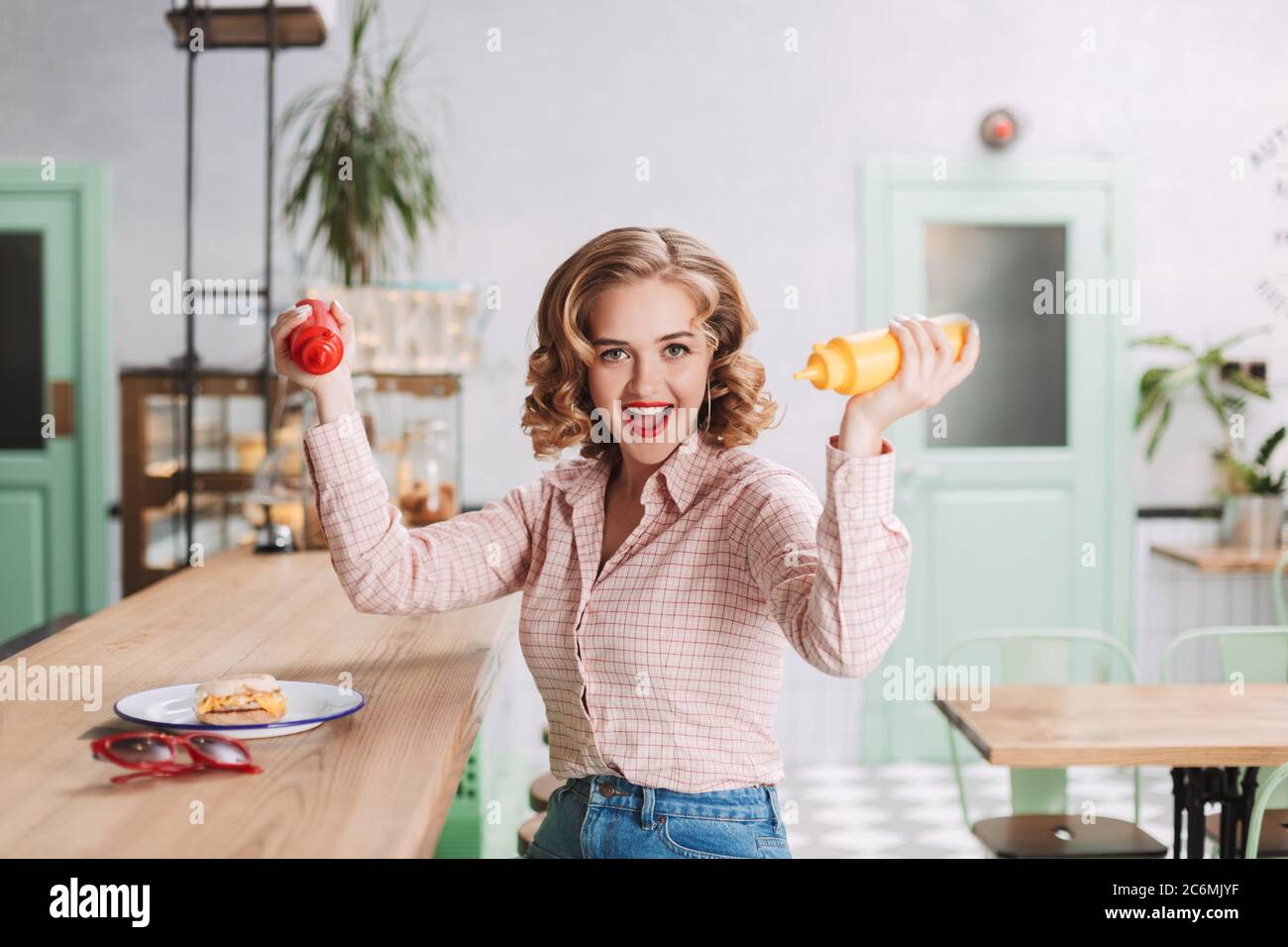 Bonita señora en camisa sentada con botellas de ketchup y mostaza en las manos y con alegría mirando en la cámara mientras pasa tiempo en el café. Foto de stock