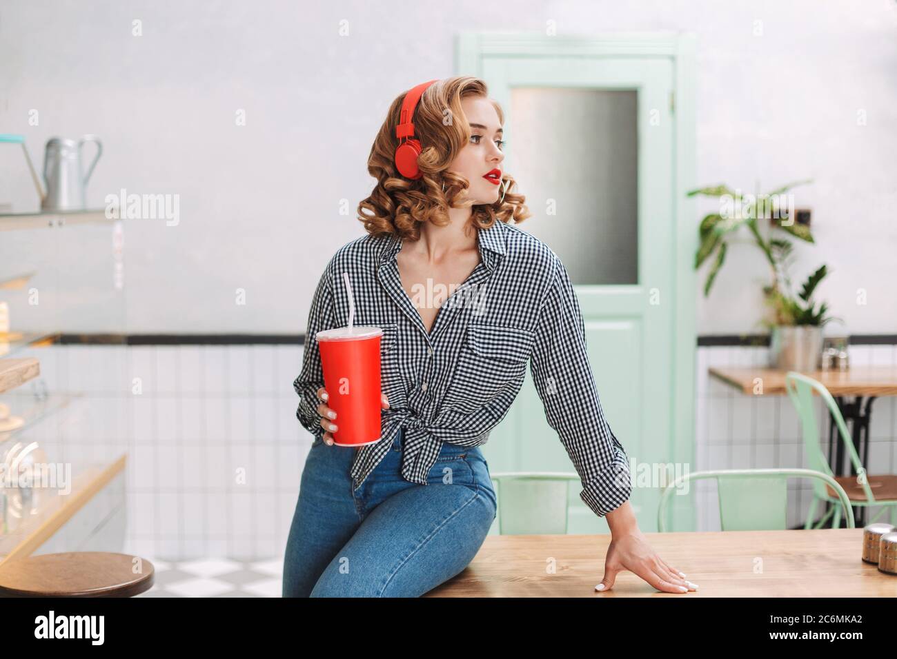 Joven y agradable dama con auriculares rojos de pie cerca de la mesa con agua de soda en la mano y mirando a un lado en el café. Foto de stock