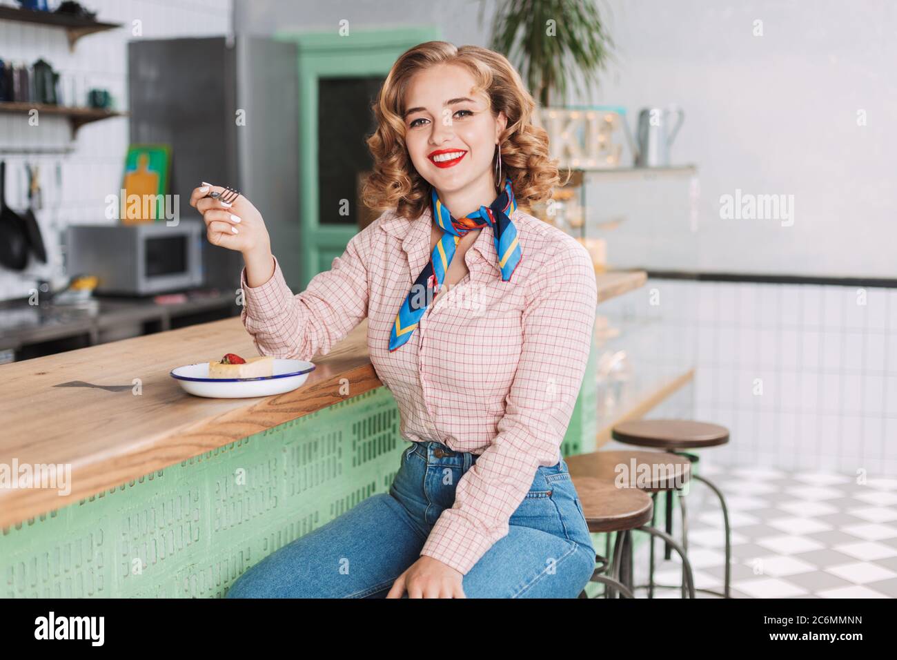 Joven hermosa señora en camisa y jeans, sentada en el mostrador del bar en el café y comiendo pastel mientras que felizmente mirando en la cámara Foto de stock