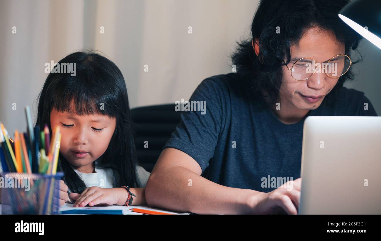 Padre usando el portátil hacer horas extras y la hija haciendo la tarea escolar en la mesa de trabajo de oficina en casa por la noche Foto de stock
