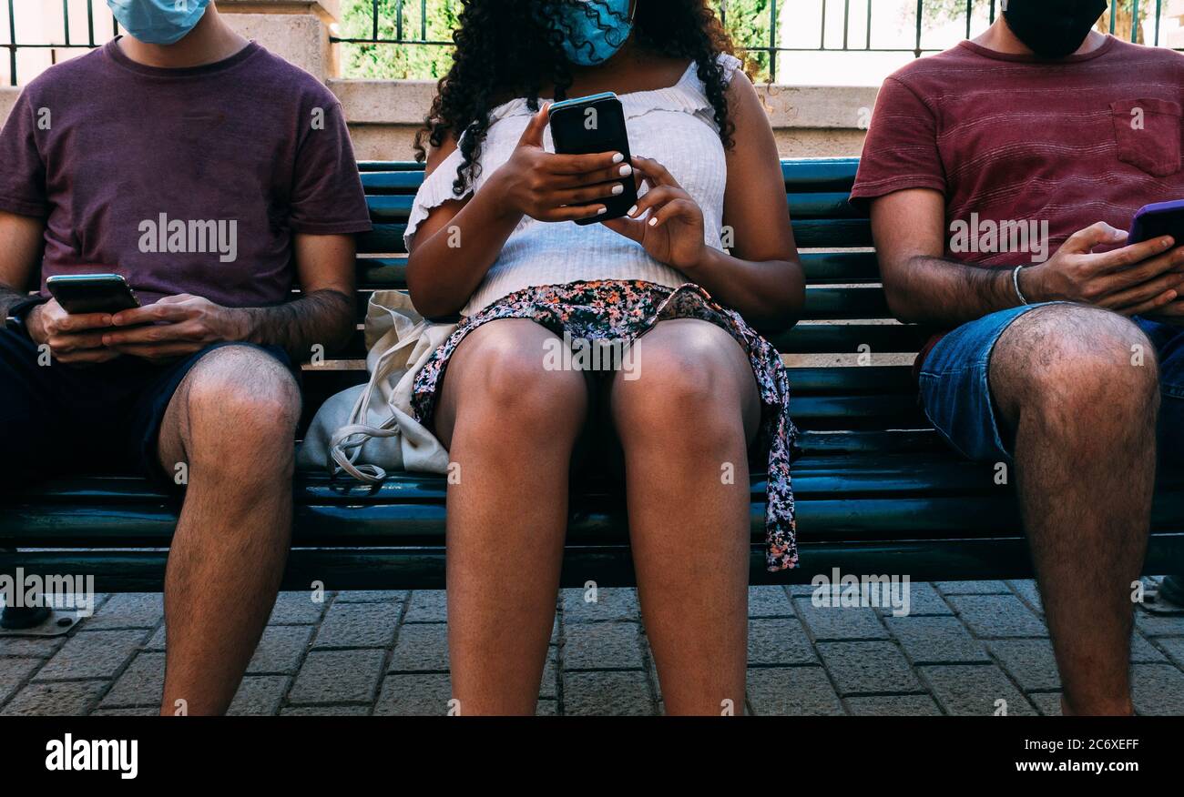 tres personas con máscara en el banco utilizando teléfono móvil, concepto de distancia social con máscara Foto de stock