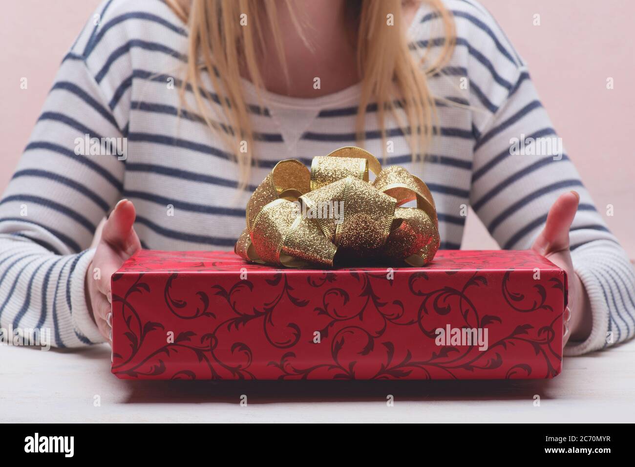 Caja de regalo para Nochebuena con patrones. Regalo y chica rubia en blusa a rayas en el fondo. Las manos femeninas sostienen la caja envuelta en papel rojo con arco de oro, Caja de regalo para Nochebuena con patrones. Regalo y chica rubia en blusa a rayas en el fondo. Las manos femeninas sostienen la caja envuelta en papel rojo con arco de oro,