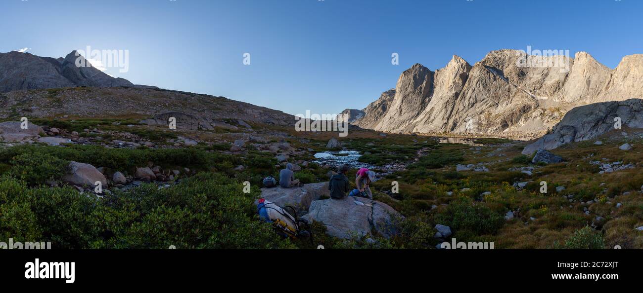 Un disparo panorámico de tres mochileros haciendo una pausa mientras se hace senderismo por la Ruta del Río viento. La luz del amanecer ilumina Ambush Peak. Foto de stock