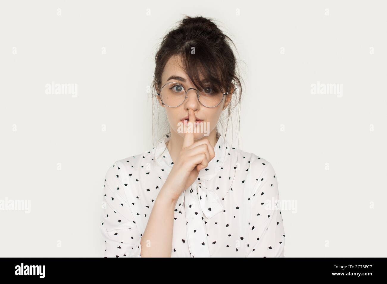 Encantadora dama morena con gafas que están gestuando el signo de silencio en una pared blanca del estudio mirando la cámara Foto de stock