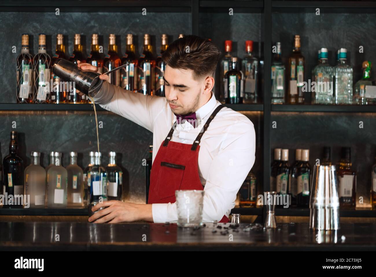 Barman profesional en uniforme haciendo muestra de su trabajo, sosteniendo  dos partes de agitador de metal en sus manos y vertiendo un cóctel,  estantes llenos de bottel Fotografía de stock - Alamy
