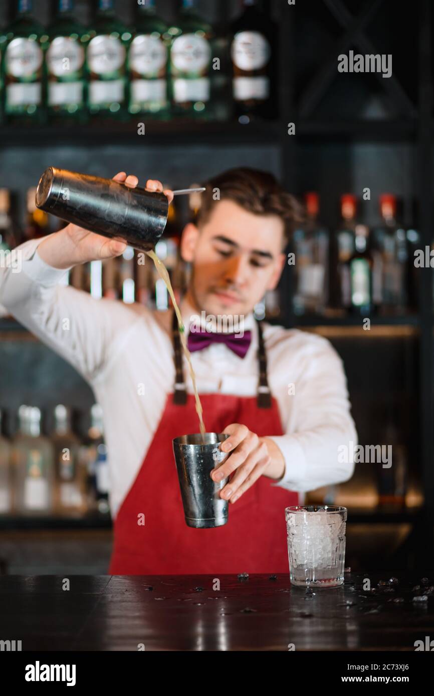 Barman profesional en uniforme haciendo muestra de su trabajo, sosteniendo  dos partes de agitador de metal en sus manos y vertiendo un cóctel,  estantes llenos de bottel Fotografía de stock - Alamy