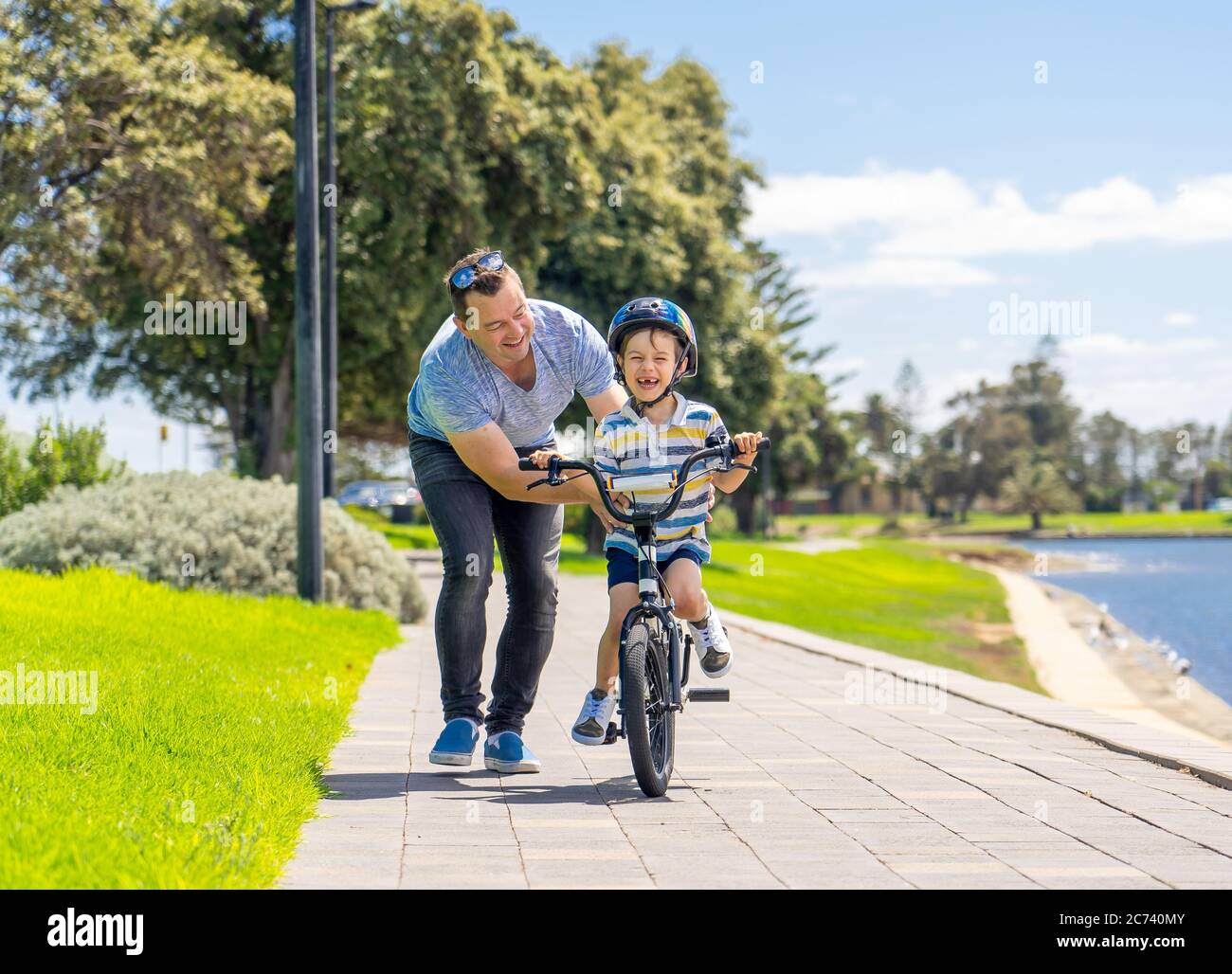 Niño aprendiendo a montar en bicicleta con su padre en el parque junto al lago. Padre e hijo se divierten juntos en las bicicletas. Feliz familia, actividades al aire libre Foto de stock