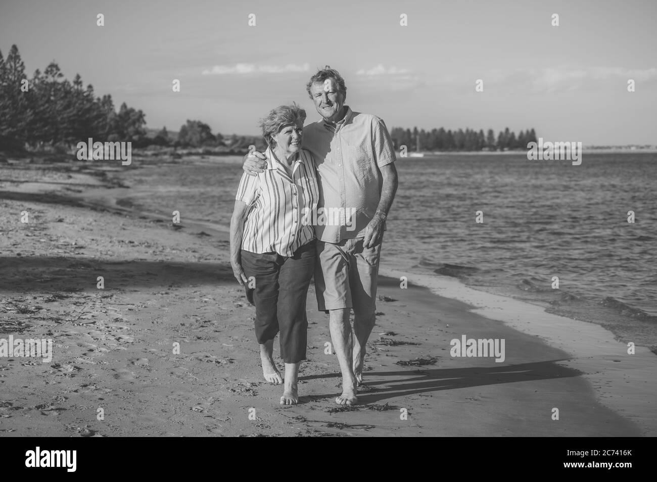 Feliz pareja mayor abrazando y sosteniendo las manos caminando en la playa vacía al atardecer. Mujer y hombre de edad activa y saludable en un paseo romántico por el mar. Foto de stock