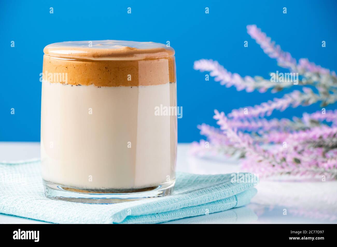Café y flores de Dalgona sobre fondo azul. Moda tendencia bebida de leche y espumas batidas dulces. Foto de stock
