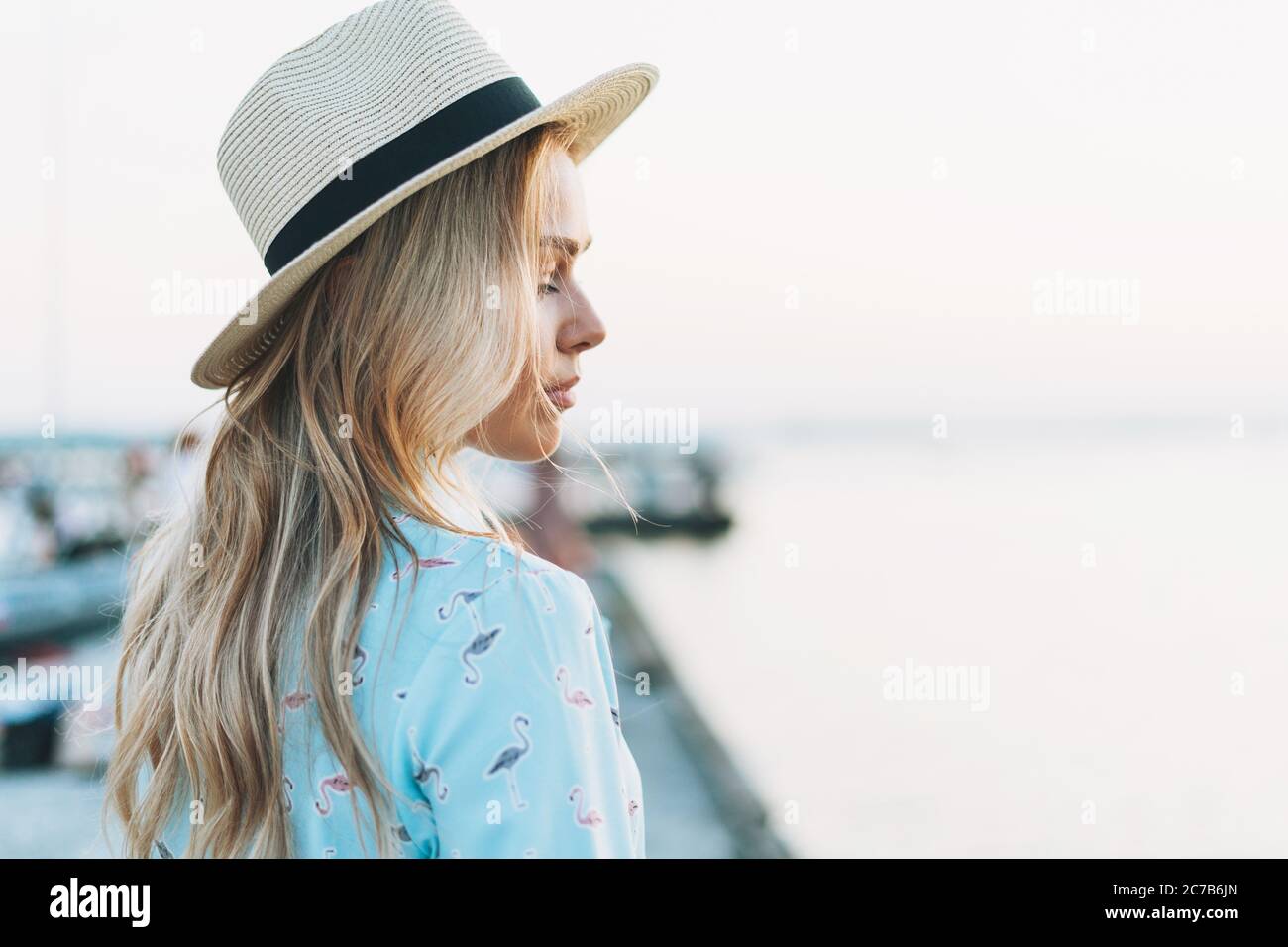 Hermosa mujer joven rubia con vestido azul y sombrero de paja en el muelle al atardecer Foto de stock