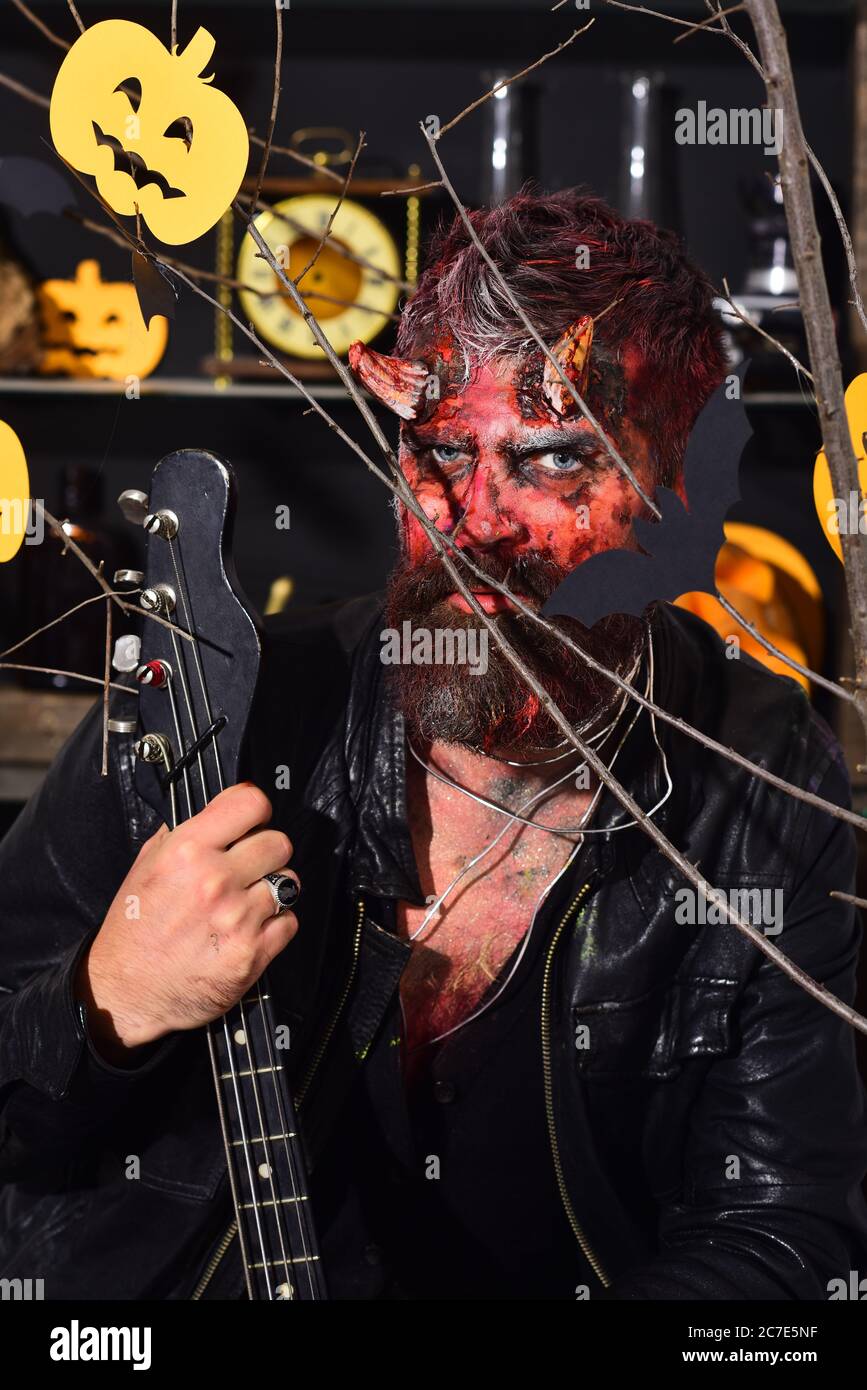 Demonio con cuernos y cara malvada sostiene guitarra eléctrica negra. Diablo  o monstruo jugando instrumento. Hombre vestido de maquillaje terrorífico  detrás de la decoración de las calabazas y murciélagos de Halloween. Disfraz