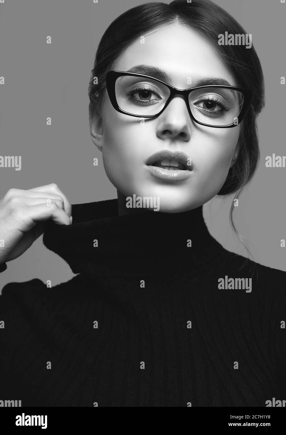 Retrato de una chica linda con un suéter de cuello turtleneck negro y gafas con estilo posando sobre fondo gris en el estudio Foto de stock