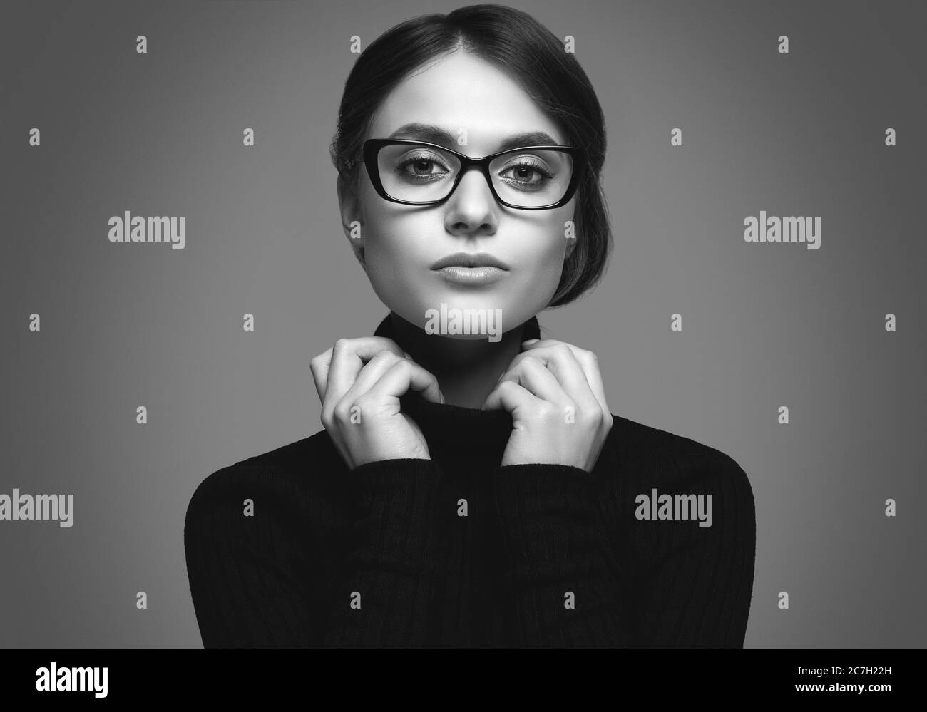 Retrato de una chica linda con un suéter de cuello turtleneck negro y gafas con estilo posando sobre fondo gris en el estudio Foto de stock