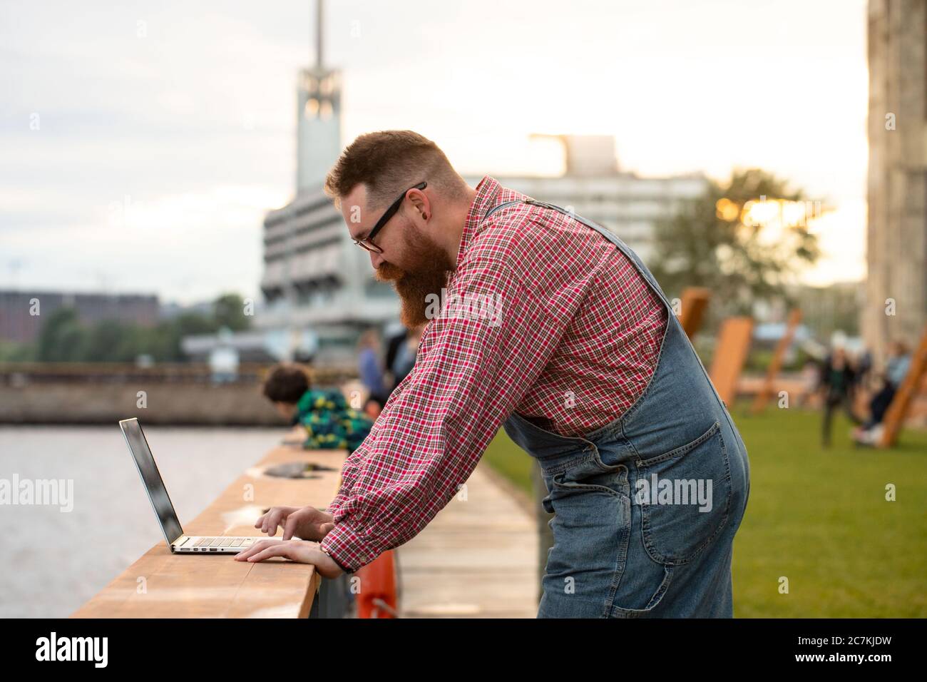 Retrato de un brutal hombre balbardo freelancer con overoles azules y  camisa de cuadros trabajando en el portátil en el terraplén al aire libre.  Trabajo a distancia, trabajo remoto Fotografía de stock -