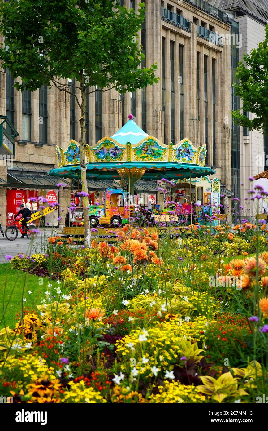 Carrusel nostálgico frente al edificio histórico de los grandes almacenes Kaufhof en Düsseldorf, con coloridas flores de verano en primer plano. Foto de stock