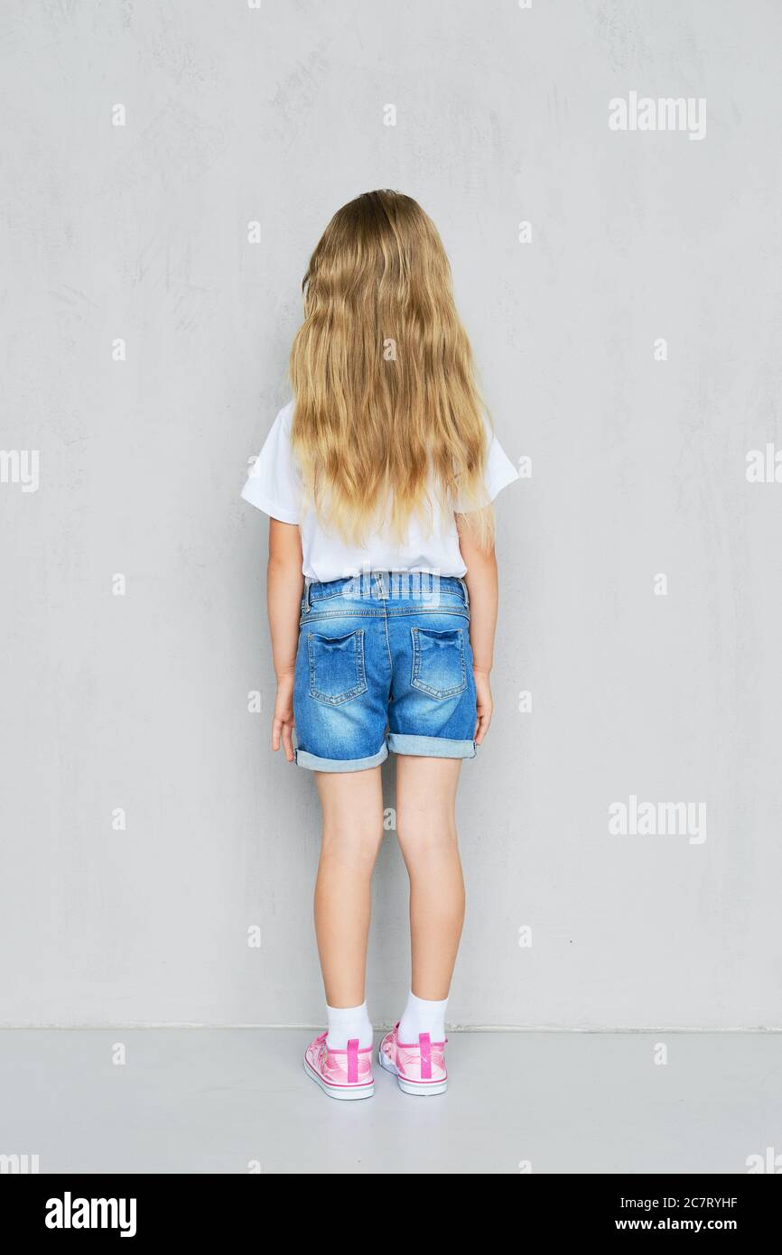 Vista posterior de niña pequeña en camiseta blanca, pantalones cortos de  vaqueros y zapatillas de deporte rosas posando cerca de la pared Fotografía  de stock - Alamy