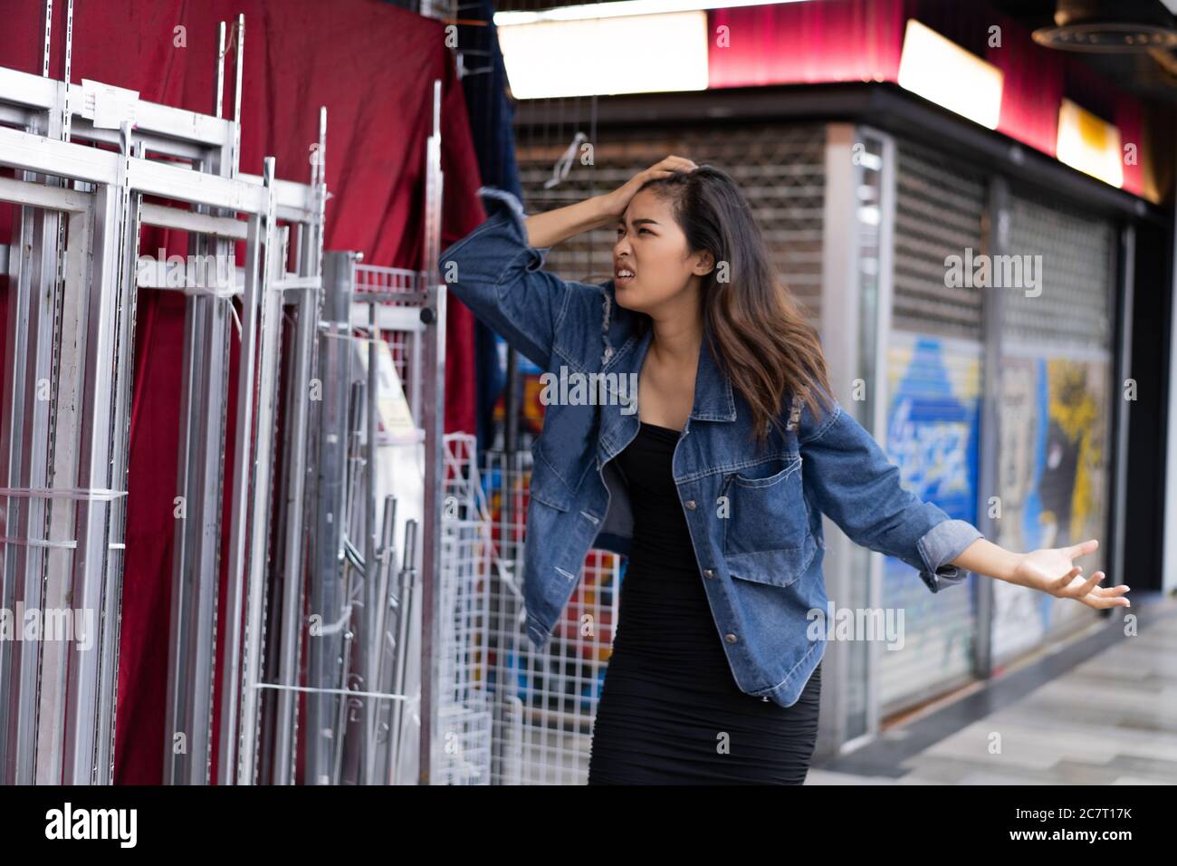 Joven mujer asiática con la mano en el cabello gestuando en la ira frente a la tienda cerrada Foto de stock