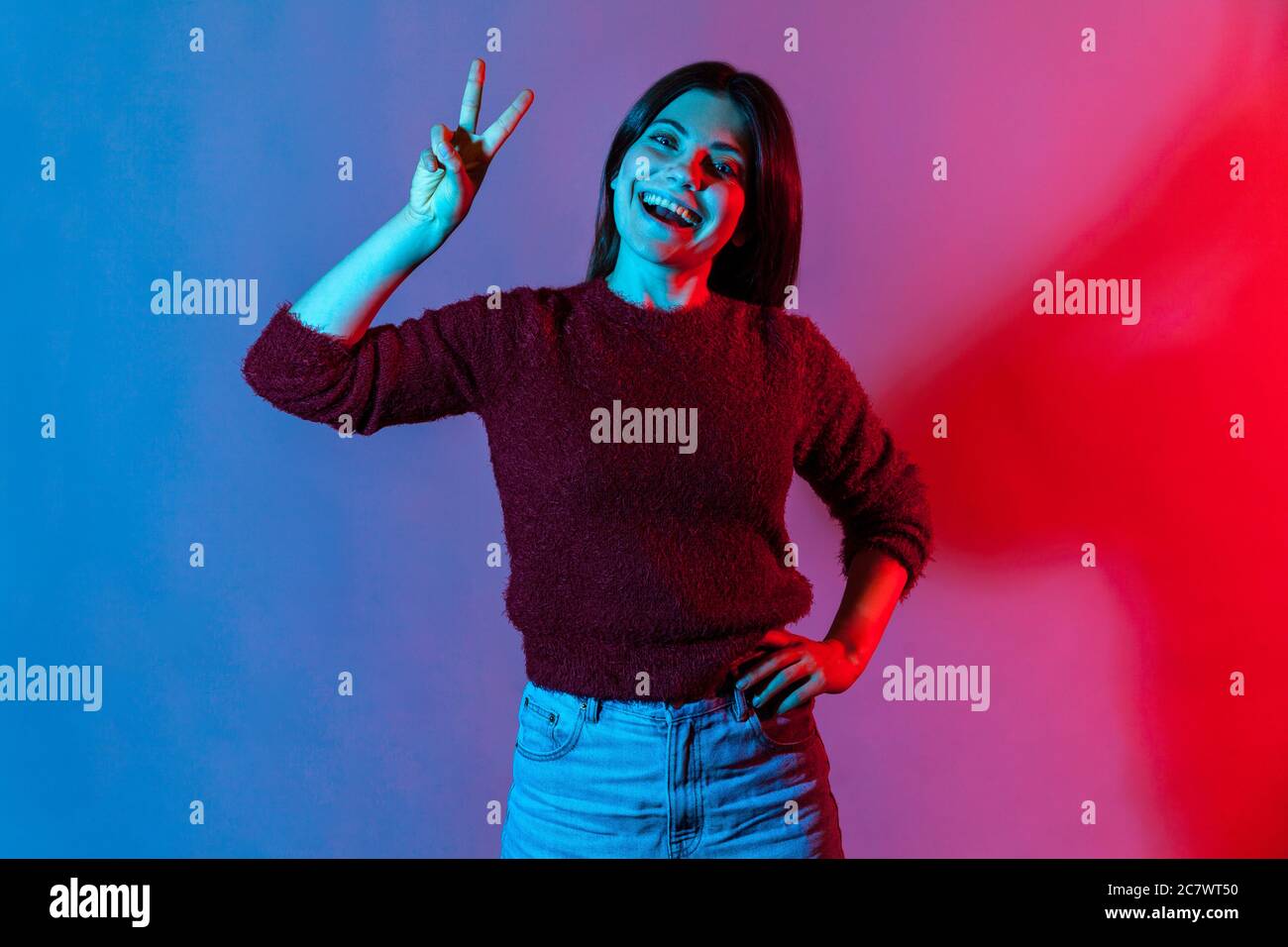 Retrato de neón ligero de mujer feliz despreocupada mostrando victoria o gesto de paz y sonriendo ampliamente, regocijándose con suerte ganando, sintiéndose optimista a Foto de stock