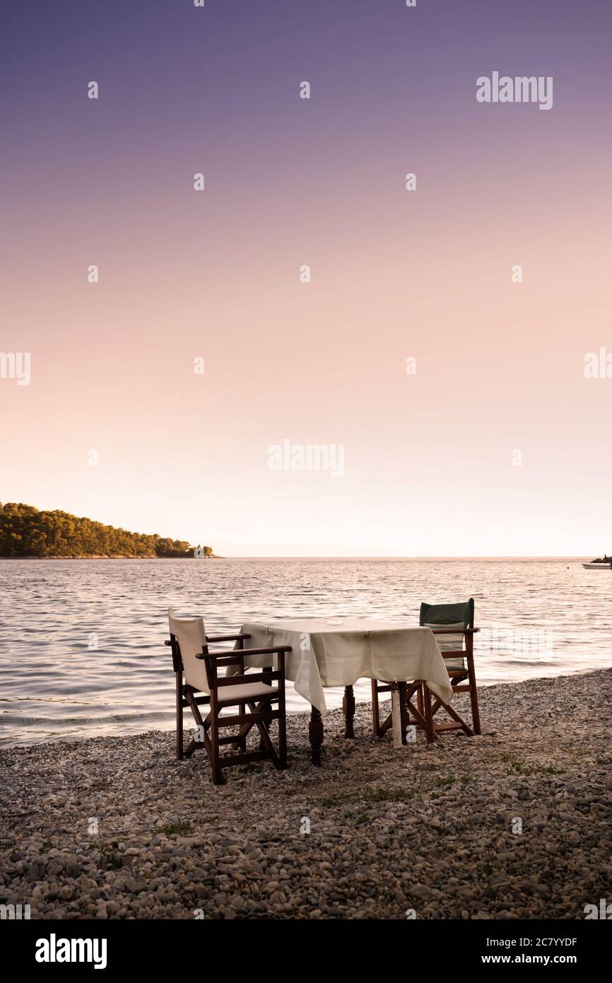 Una mesa y dos sillas se encuentran en la playa cerca del mar Mediterráneo, bajo el cálido sol de la noche, esperando una cena romántica. Foto de stock