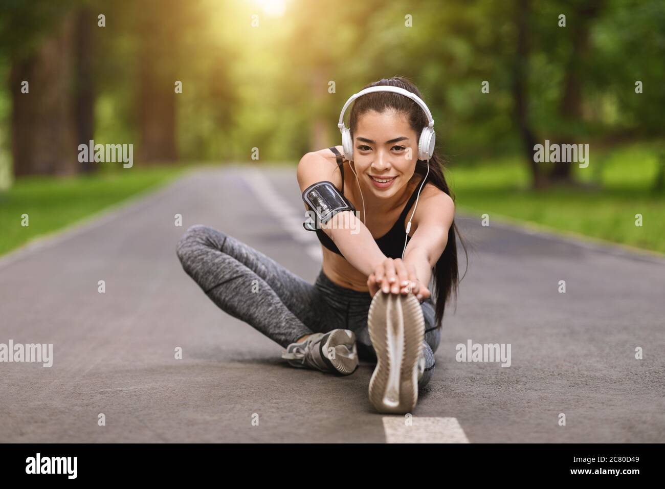 Entrenamiento de fitness al aire libre. Chica asiática sonriente estirándose en el parque y escuchando música Foto de stock