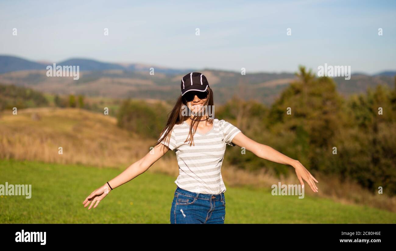 Hermosa mujer sin preocupaciones en gafas de sol, gorra y pantalones cortos  pantalones cortos de jeans caminar en el campo de verano con las montañas  en el fondo Fotografía de stock -