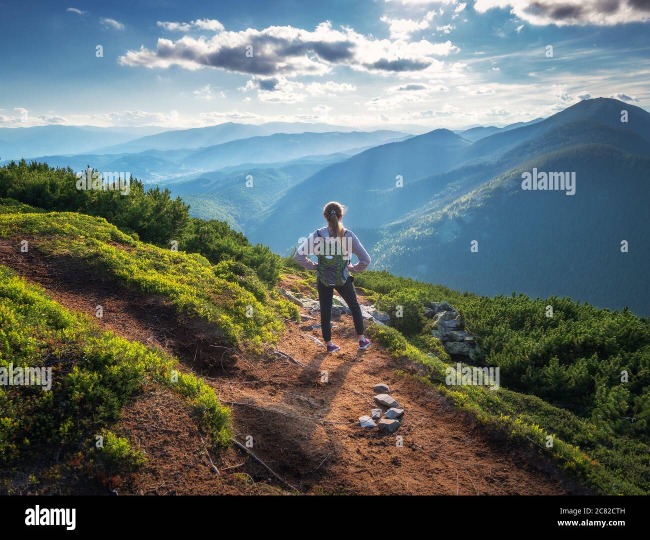 Montañas y mujer joven de pie con mochila en el camino Foto de stock