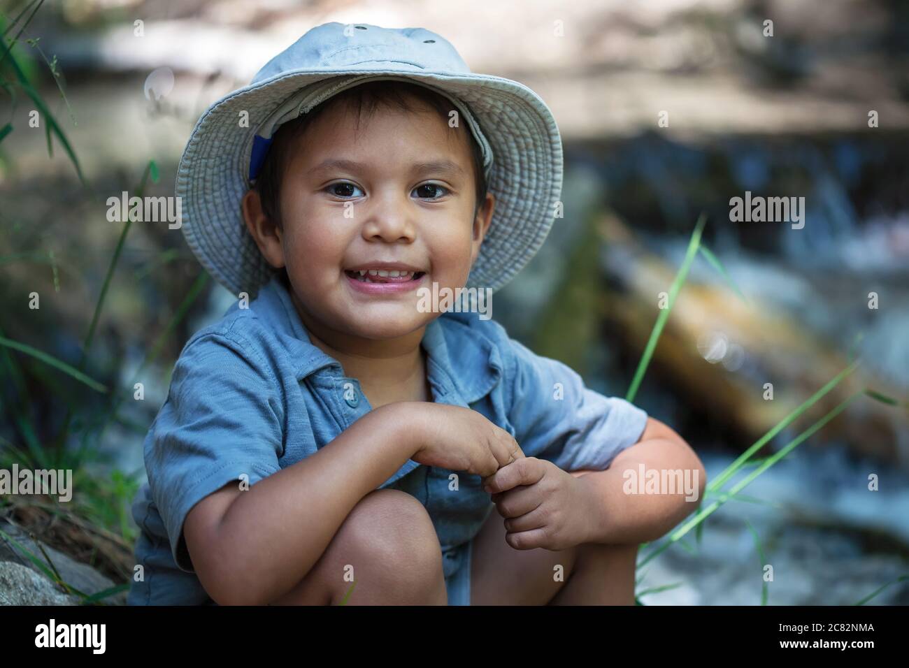Un niño pequeño con su sombrero de pesca, sentado junto al borde del río durante el verano. Foto de stock