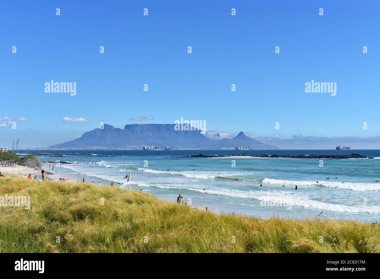 Table Mountain, Devils Peak y Lions Head vistos desde la playa de Bloubergstrand desde el otro lado de Table Bay. Se puede ver a la gente en la playa y en el agua. Foto de stock