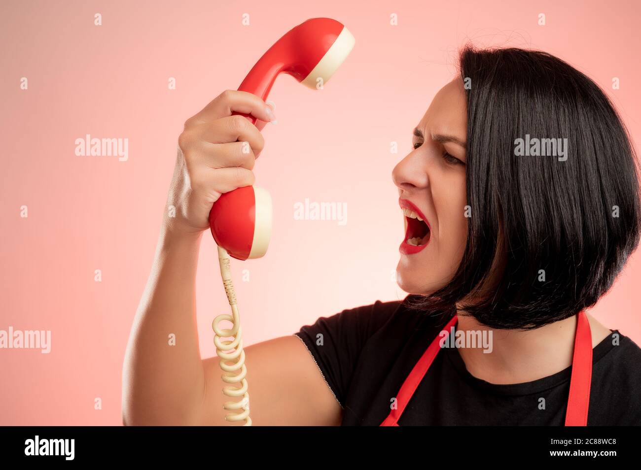 Mujer empleada en el supermercado con delantal rojo y camiseta negra, gritó en el receptor del teléfono aislado sobre fondo rojo Foto de stock