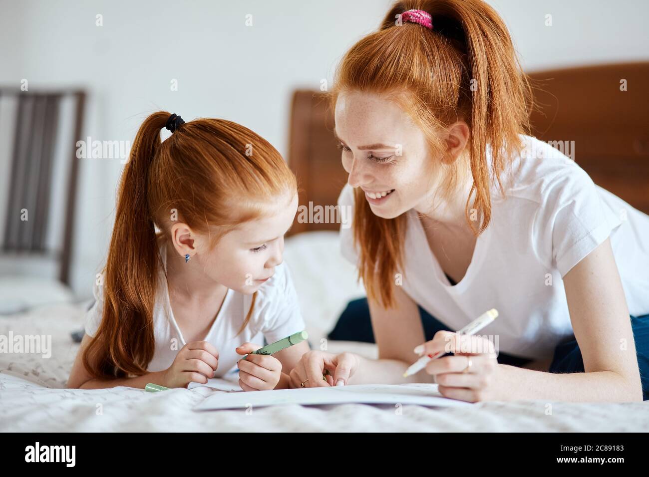 Bonita niña con cola de caballo de jengibre tumbada en la cama con su madre y usando lápices de colores para hacer la tarjeta de Navidad, interior de la acogedora sala de estar o Foto de stock