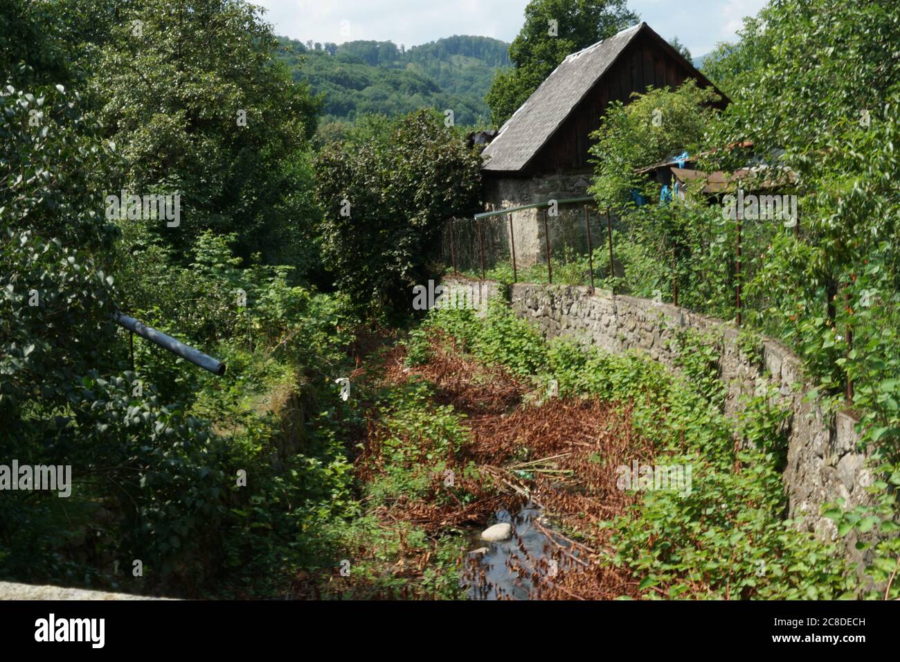 Una antigua casa de pueblo abandonada casi perdida entre la vegetación en crecimiento. Un muro de piedra o barrera lo protege contra el arroyo en un pueblo. Foto de stock