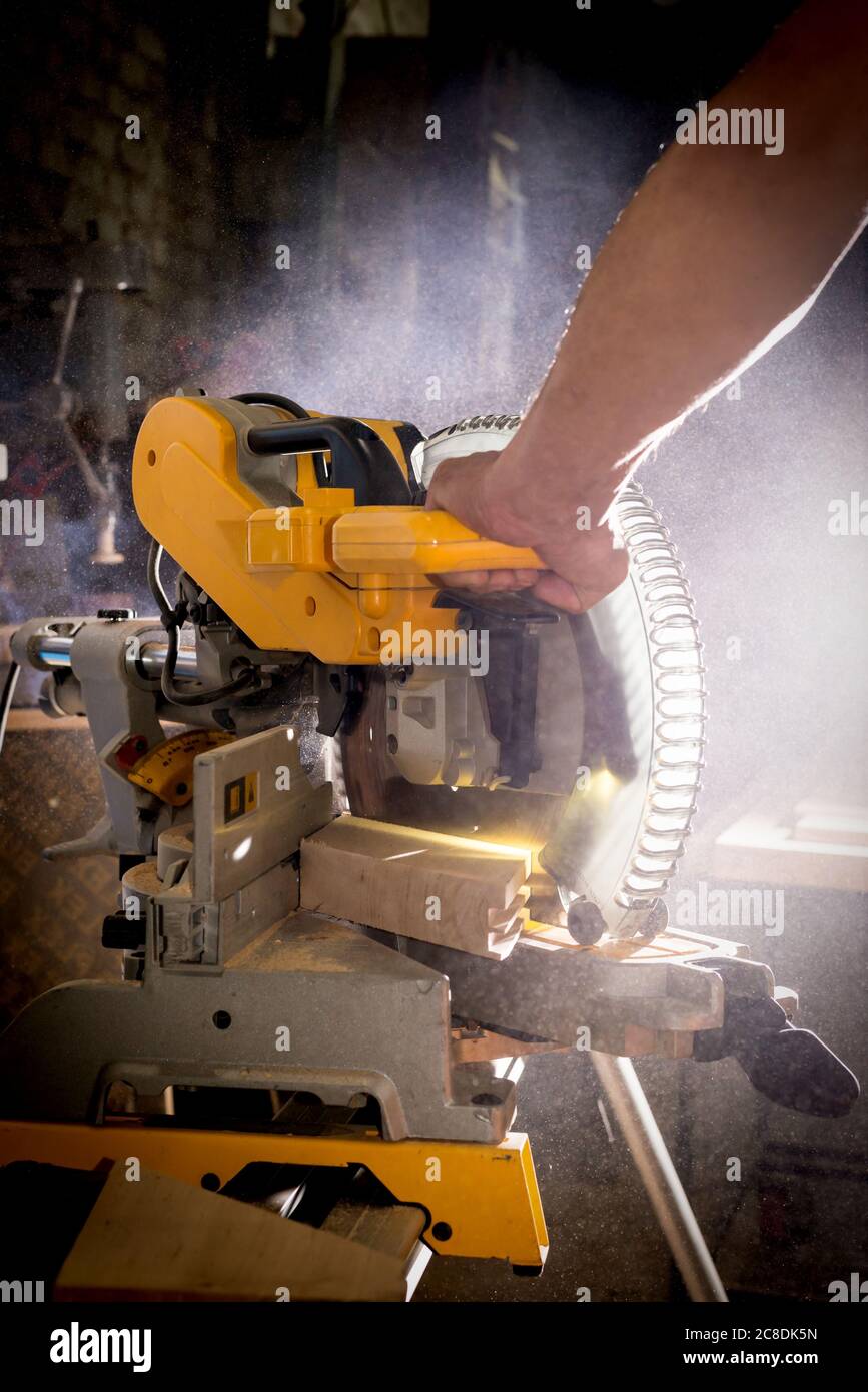 Carpenter trabajando con la miter circular de poder para cortar la tabla de madera contra el fondo oscuro. Foto de stock