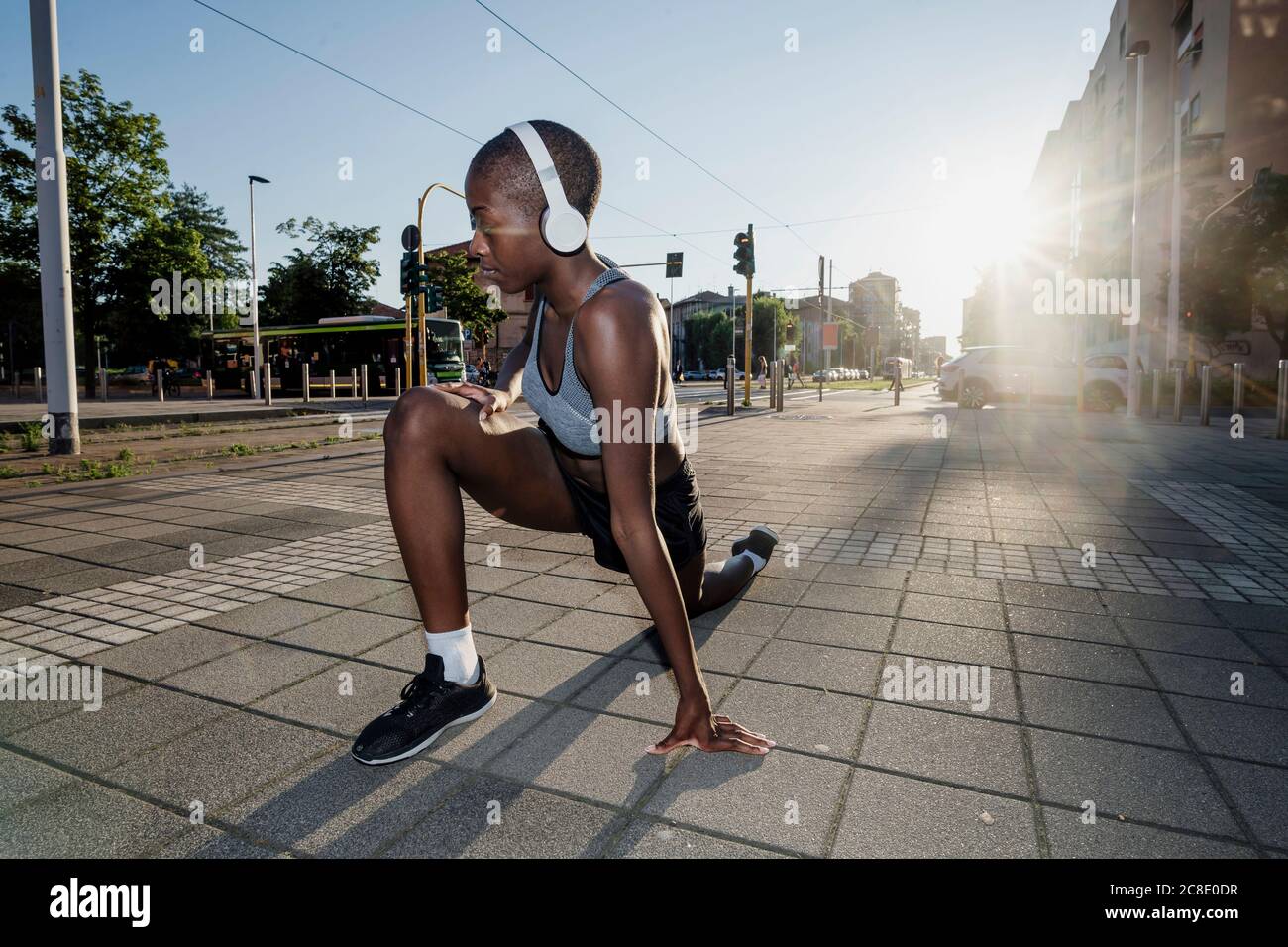 Mujer con cabeza afeitada escuchando música a través de los auriculares mientras se estira piernas en la calle en la ciudad Foto de stock