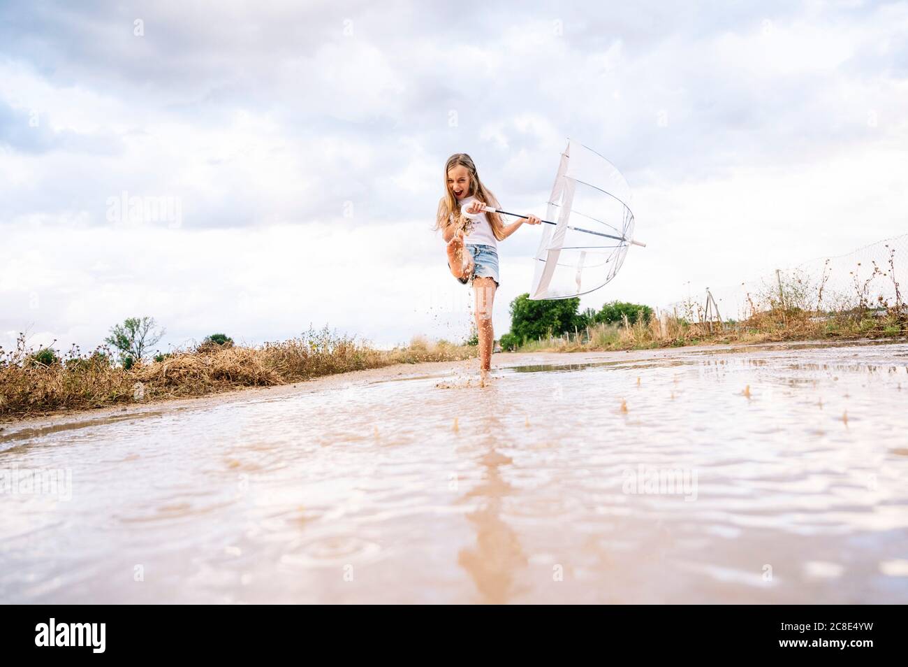 Chica alegre chapoteando agua en charco mientras sostiene el paraguas contra cielo nublado Foto de stock