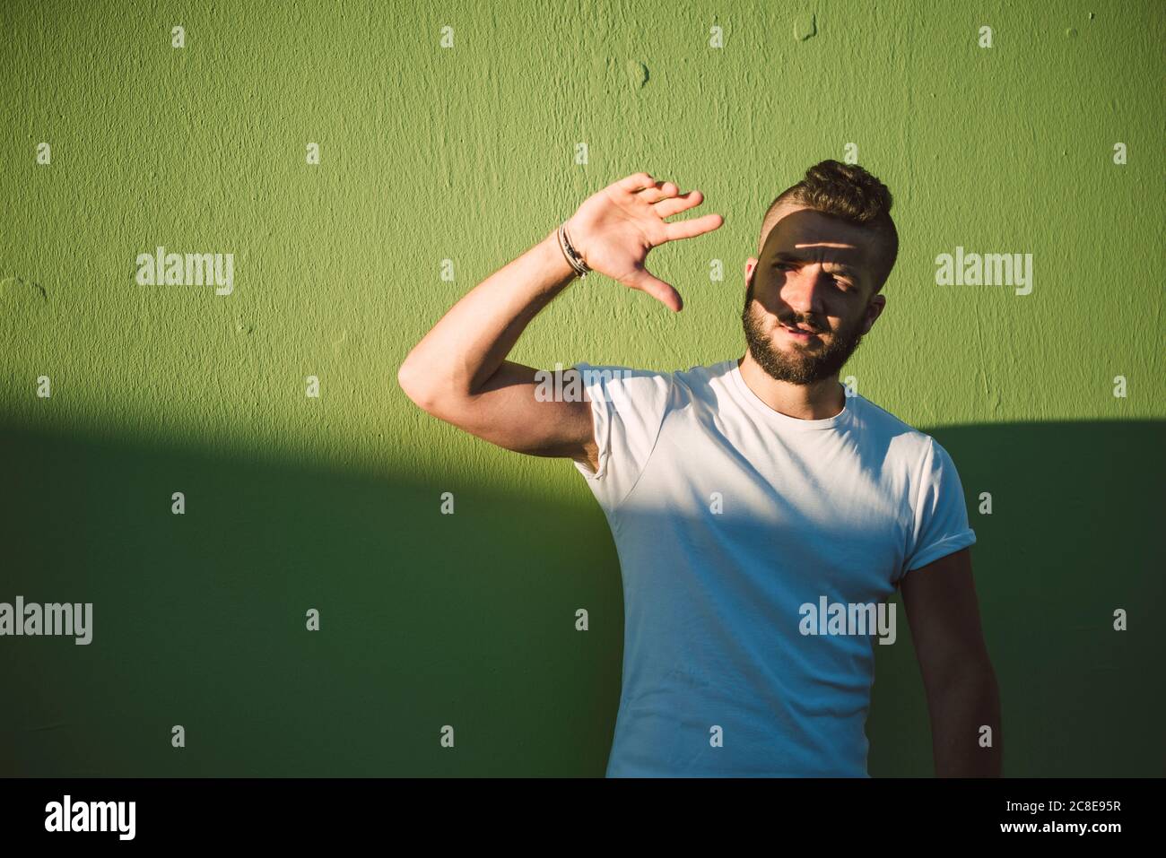 Hombre joven y guapo que protege los ojos mientras está de pie contra la pared verde Foto de stock