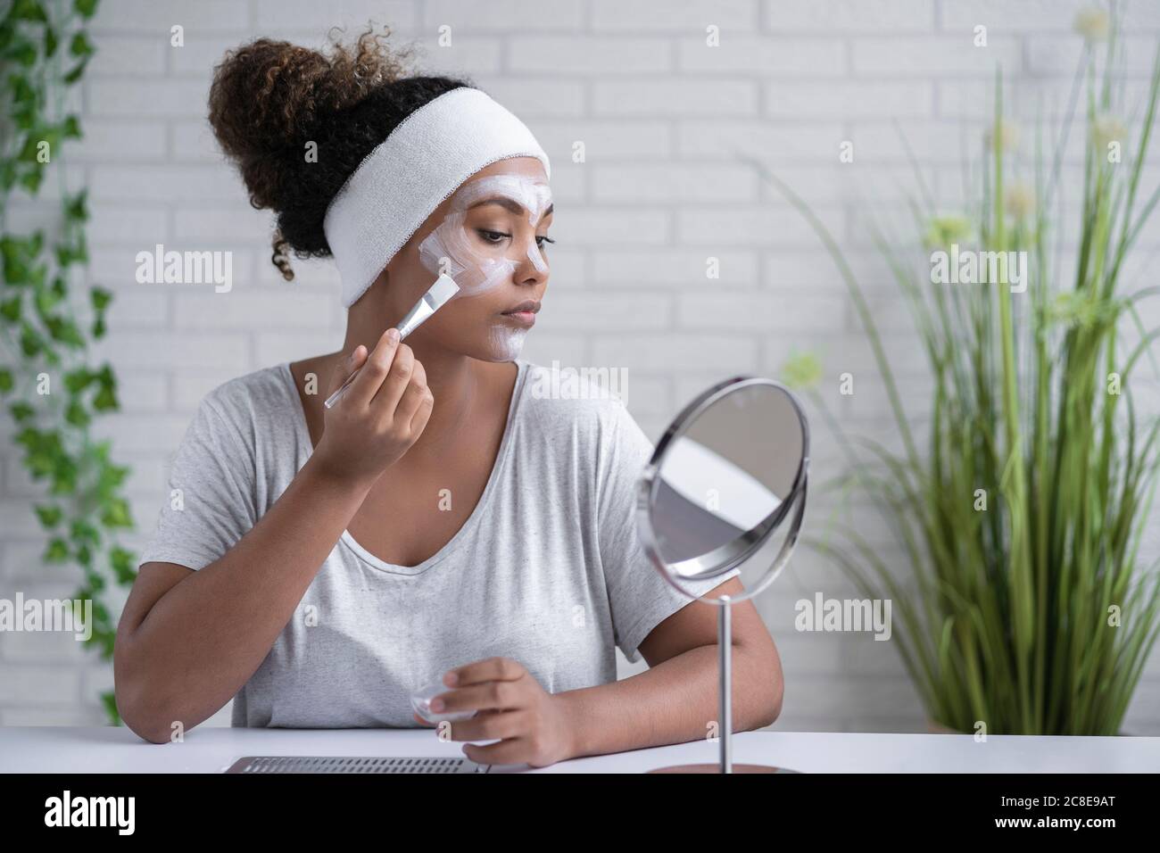 Mujer que usa cinta para la cabeza aplicando máscara facial mientras mira en el espejo en casa Foto de stock