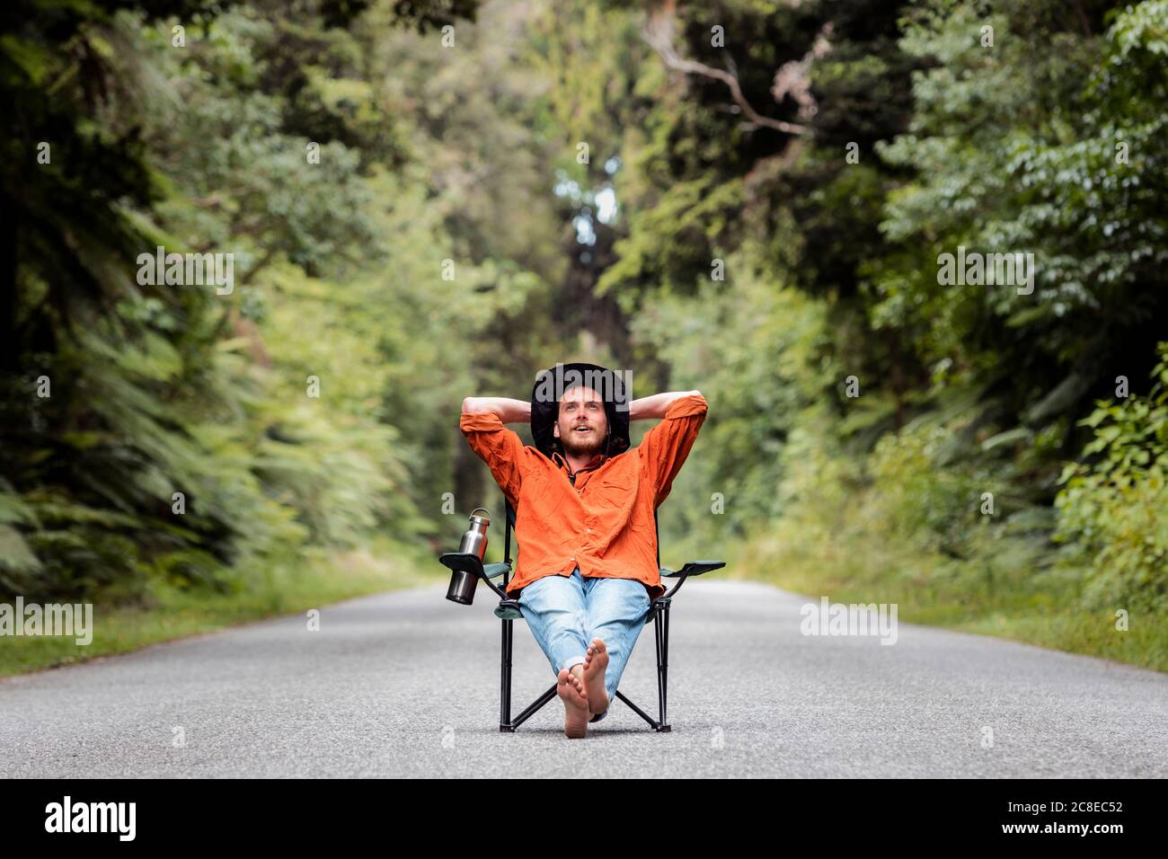 Hombre pensativo con las manos detrás de la cabeza sentado en la silla en la carretera de campo en medio de los árboles Foto de stock