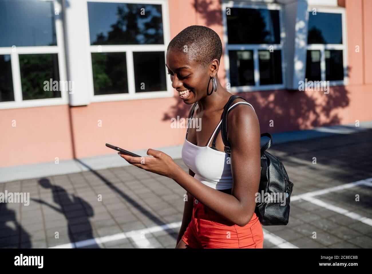 Alegre mujer joven con cabeza afeitada usando el teléfono inteligente mientras caminar en la acera de la ciudad Foto de stock